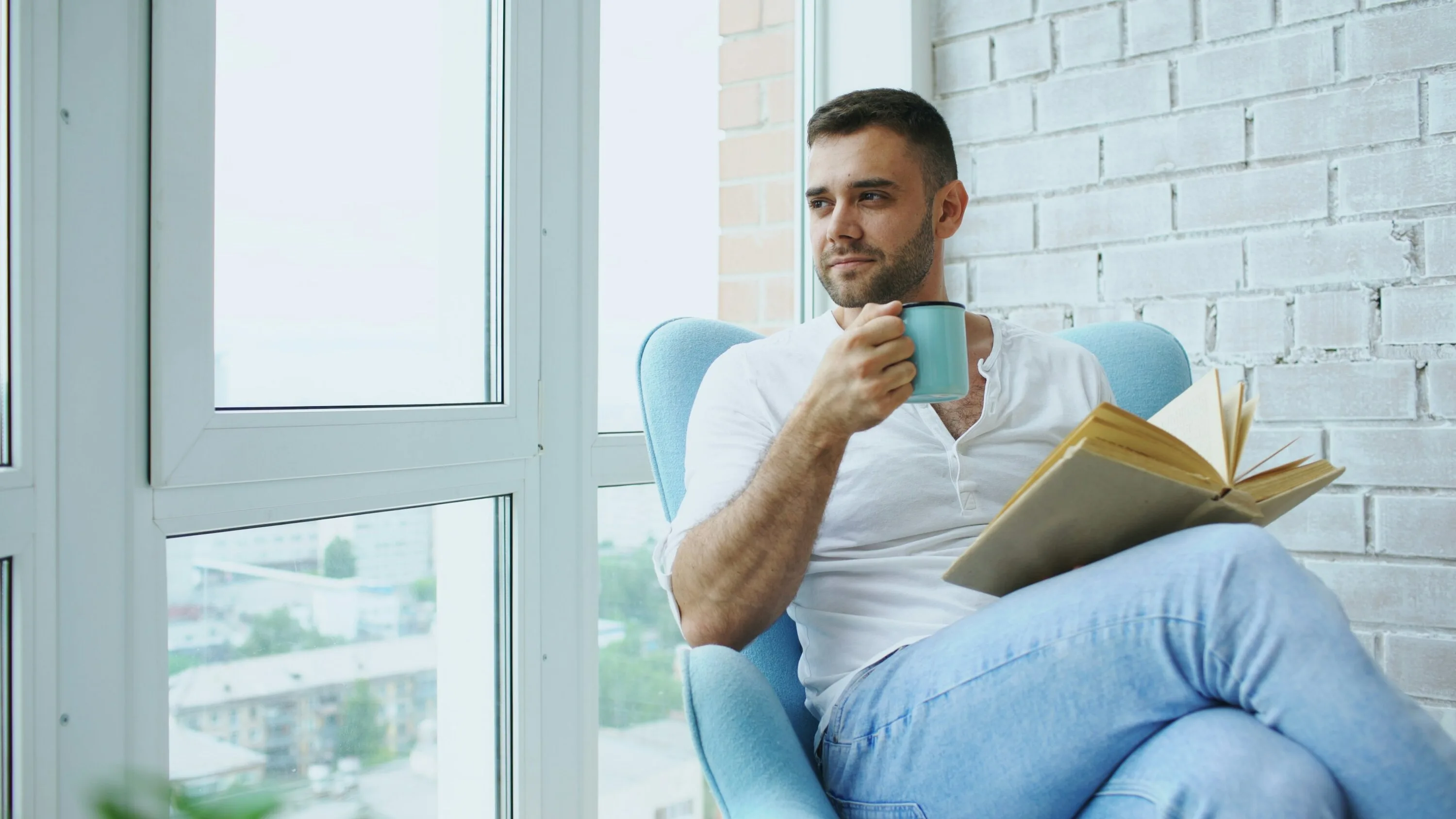 A man reading a book with a cup of tea by a bright window